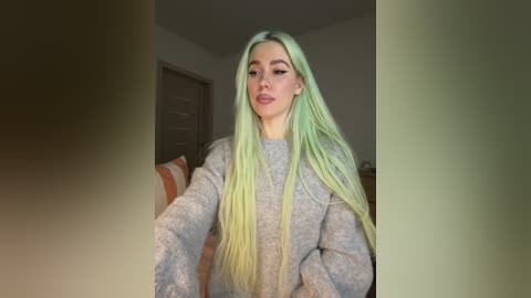 Video of a young woman with long, pastel green hair, wearing a gray, textured sweater, sitting on a striped couch, indoors, dim lighting.