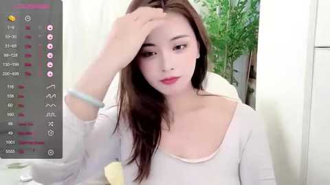 Media: Video of a young East Asian woman with fair skin and long brown hair, wearing a white long-sleeve top, sitting indoors. She is resting her head on her hand, looking tired. A digital interface with health metrics and icons is displayed in the background.