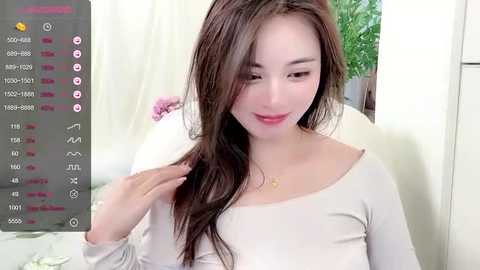 Video of a smiling, fair-skinned East Asian woman with long brown hair, wearing a beige top, adjusting her hair in a home setting.