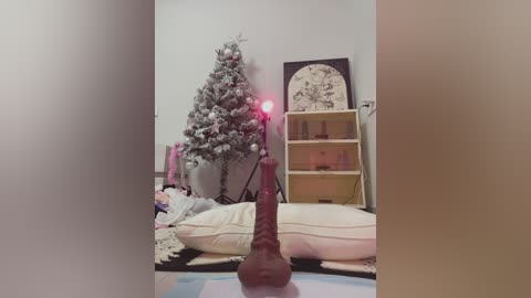 Media: Video of a cozy, dimly lit room with a decorated Christmas tree, a large wooden dildo, and a beige bookshelf, featuring a soft, pastel color palette.