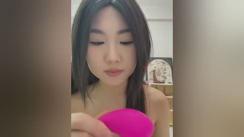 Media: A video of a young Asian woman with straight, long black hair, wearing pink lipstick, holding a bright pink vibrator, standing in a dimly lit room with wooden furniture.