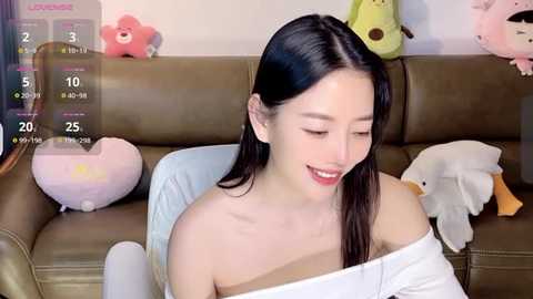 Media: Video of an East Asian woman with long black hair, light skin, and red lipstick, wearing a white off-shoulder top, sitting on a green leather couch with stuffed toys, including a pink bunny and a bear, in a cozy room.