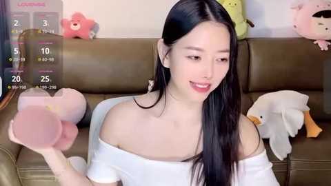 Media: Video of a young East Asian woman with long black hair, fair skin, and an off-shoulder white top, smiling, holding stuffed animals in a cozy living room with a brown couch and plush toys.