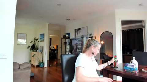 Media: Video of a blonde woman in a white t-shirt working at a desk in a cozy, modern living room with light walls, wooden floor, and plants.