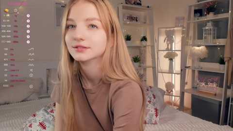 Media: Video of a young Caucasian woman with long blonde hair, fair skin, and light makeup, wearing a beige top, sitting on a bed with floral-patterned sheets in a cozy, well-lit bedroom.