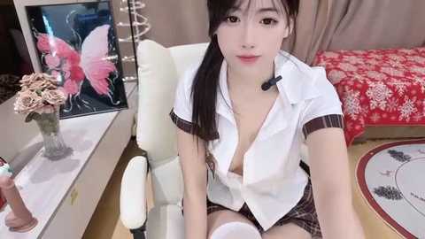 Media: A video of an Asian woman with long black hair in a white, low-cut blouse, plaid skirt, and white thigh-high socks, sitting in a modern, beige office chair, surrounded by a floral-patterned bed and a pink butterfly painting.