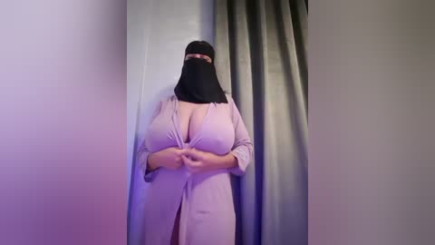 Media: Video of a plus-sized woman in a light pink, low-cut, button-up dress, wearing a black niqab with a pink headband, standing against a white wall with a gray curtain.