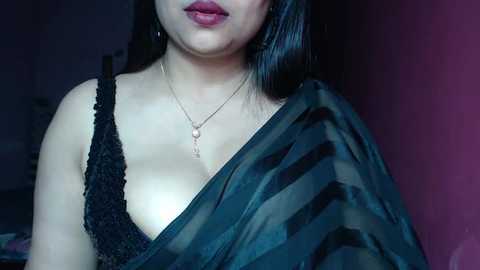 Media: Video of a woman with light skin and black hair, wearing a black lace bra and a dark shawl. She has a small, round pendant necklace. Background is a dark, blurred room.