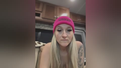 Media: Video of a blonde woman with a pink beanie and tattoos, topless, leaning forward, in a kitchen.