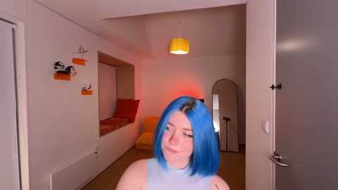 Video of a young woman with bright blue hair, standing in a modern, minimalist bedroom with white walls, a red accent light, and a yellow pendant lamp.