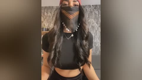 Media: Video of a young woman with long, wavy dark hair, wearing a black crop top, black face mask with red and orange geometric patterns, and a red beanie. Background features a textured, grey stone wall.