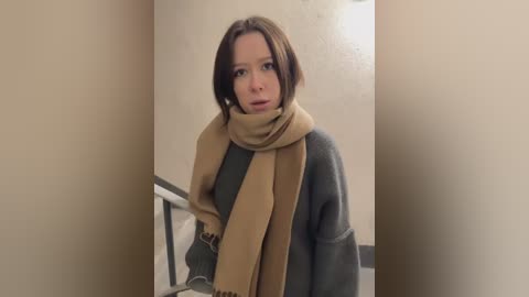 Media: Video of a woman with shoulder-length brown hair, wearing a beige scarf and a gray sweater, standing in a beige hallway with a metal railing.