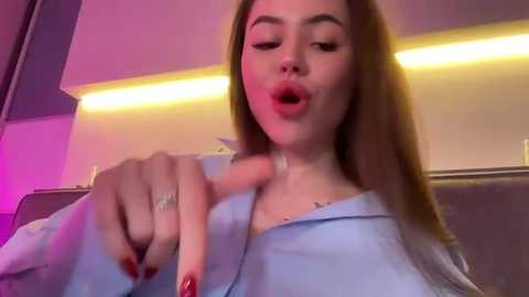 Media: Video of a light-skinned woman with long brown hair, wearing a light blue shirt, licking her finger, in a dimly lit room with purple and yellow lights.