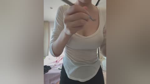 Media: A video of a woman with light skin, wearing a white, long-sleeve top and black pants, standing in a bedroom with a bed and curtains in the background.