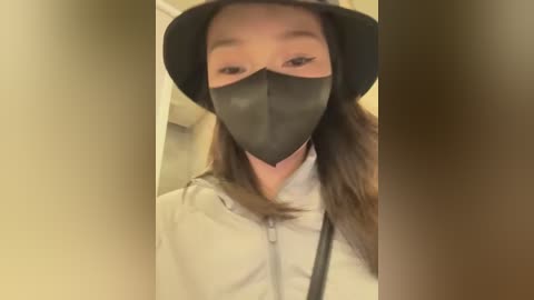 Media: A video of an Asian woman with light skin, wearing a black face mask and a black hat, standing in a beige hallway.