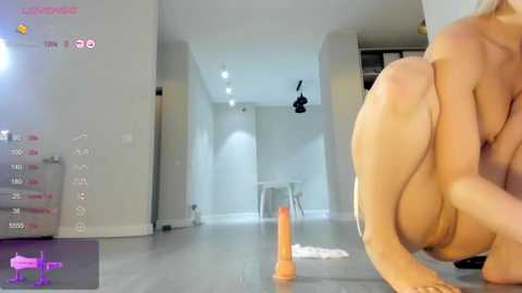 Media: Video of a nude, light-skinned woman in a modern, minimalist apartment. She is squatting on the floor, holding an orange dildo. The room has white walls and a wooden floor.