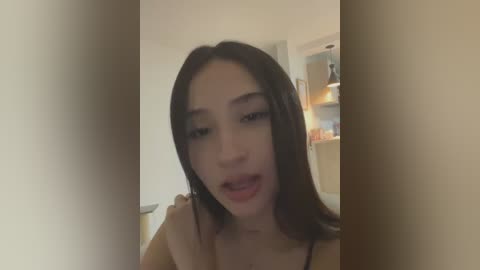 Media: Video of an Asian woman with long black hair and light skin, wearing a brown top, leaning forward in a dimly lit room with a beige wall and shelves.