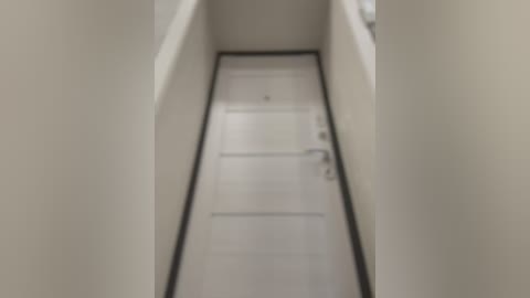 Media: Video of a narrow, dimly lit hallway with beige walls and a white door with a silver handle at the end. The floor is covered in smooth, light-colored tiles, giving a minimalistic and clean appearance.