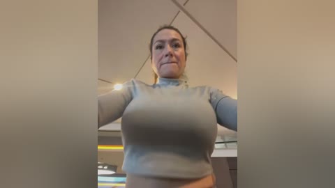 Media: A video of a middle-aged woman with fair skin and brown hair tied back, wearing a tight, light gray long-sleeve top that accentuates her large breasts. She stands in a modern kitchen with stainless steel appliances.