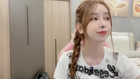 Media: Video of a young Asian woman with light skin and long brown hair in braids, wearing a white T-shirt with black text, sitting in a modern room with light-colored wooden furniture and a pink chair.