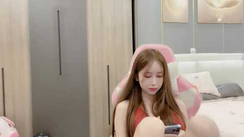 Media: Video of a young Asian woman with long, straight brown hair, sitting in a pink gaming chair, wearing a red top, focusing on a smartphone. The background features a modern bedroom with light-colored wooden wardrobes and a white bed with pillows.