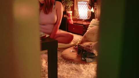 A video of a woman in a cozy, dimly lit room, seated on a fur rug, wearing a pink nightgown. A lamp and potted plants are visible in the background.