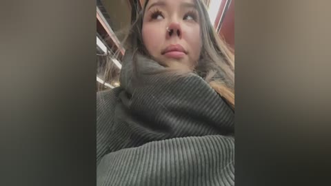 Media: Video of a young woman with fair skin and long brown hair, wrapped in a grey, ribbed knit scarf, looking up thoughtfully in a dimly lit room.