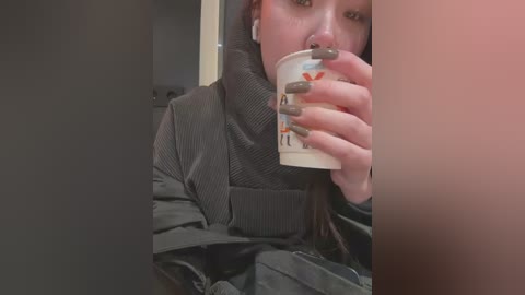 Media: Video of a woman with light skin, fair complexion, and long dark hair, wearing a black puffer jacket and grey gloves, holding a white coffee cup with a red logo. Background includes a beige wall and a black door.