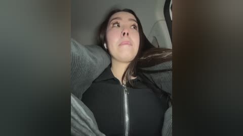 Media: Video of a young woman with light skin and dark hair, lying back in a car seat, looking up. She wears a black hoodie and has headphones on, with a muted, dark interior background.