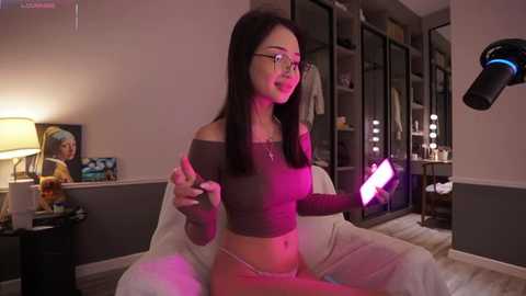 Media: Video of an Asian woman with long black hair, wearing glasses and a crop top, sitting on a white chair in a dimly-lit bedroom, holding a glowing pink remote.