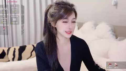 Media: Video of a smiling young East Asian woman with long, straight black hair in a high ponytail, wearing a black, low-cut top, in a modern, minimalist bedroom.