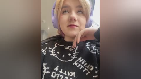 Media: Video of a young woman with fair skin and light brown hair wearing purple headphones, a black hoodie with white cat illustrations, and a contemplative expression.