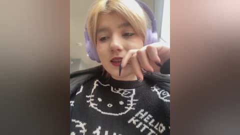 Media: Video of a young woman with light skin and blonde hair wearing purple headphones and a black sweater with white cat and \"Hello Kitty\" designs. She has a neutral expression, and the background is blurred.