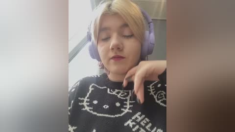 Video of a young woman with short blonde hair, wearing purple headphones and a black sweater with white cat designs. She has a neutral expression, one hand resting on her chin.