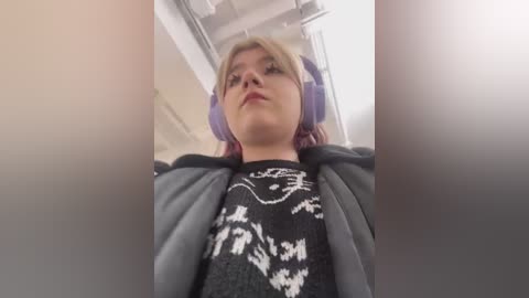 Video of a young woman with short blonde hair wearing a black sweater with white graphic design, purple headphones, and a grey jacket. She stands in a brightly lit room with a white ceiling and fluorescent lights.