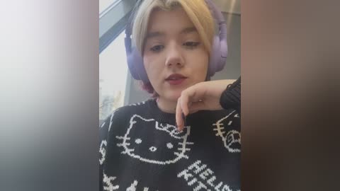 Media: Video of a young blonde woman with light skin, wearing large purple headphones and a black sweater featuring white cat and text patterns. She rests her chin on her hand, looking introspective. Background includes a blurred window.