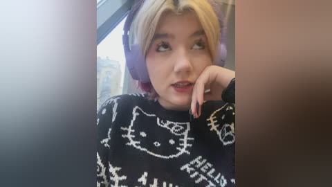 Video of a young woman with blonde hair and fair skin, wearing a black sweater with a white cartoon cat design, seated in a car, looking contemplative.