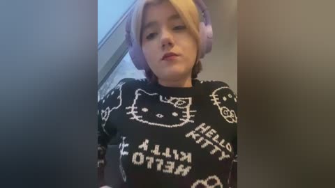 Video of a young woman with light skin, blonde hair, wearing large pink headphones, black Hello Kitty sweater, and red lipstick, looking slightly off-camera, in a dimly lit room.