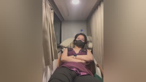 A video of a woman with dark hair and a black mask, wearing a purple top and black pants, lying on a hospital bed with beige curtains, dim lighting.