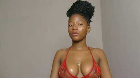 Video of a young African woman with dark skin, wearing a red lace bra with a heart pendant, her hair styled in a high bun, against a plain beige background.