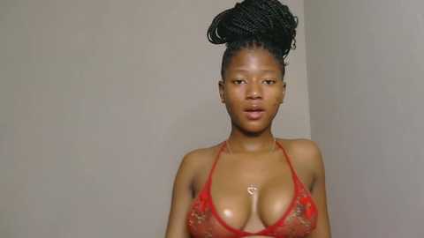 Video of a dark-skinned African woman with a curvy physique, wearing a sheer, red lace bralette that accentuates her large breasts. Her hair is styled in an updo. The background is plain beige.