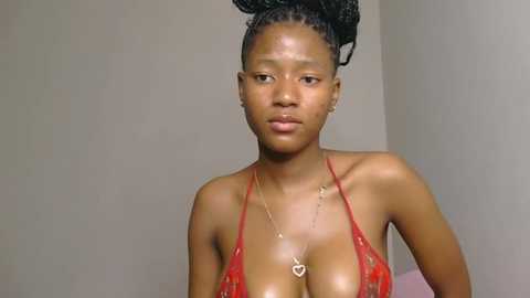 Video of a dark-skinned African woman with medium-sized breasts, wearing a red lace bra and heart pendant necklace, against a plain beige wall background.