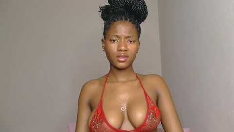 Video of a young African woman with medium-brown skin and a slender physique, wearing a red lace bra that reveals cleavage, her hair styled in a high, intricate bun. She stands against a plain beige wall, expression neutral.