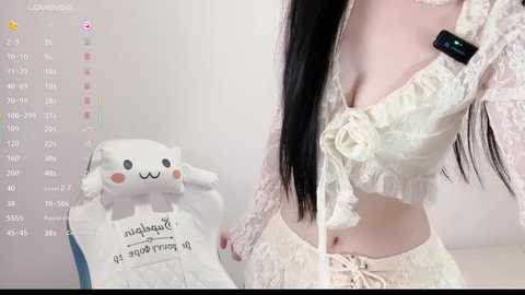 Media: Video of a slim, pale-skinned woman with long black hair, wearing a lace-up, off-the-shoulder white crop top and matching skirt, standing next to a plush cat calendar.