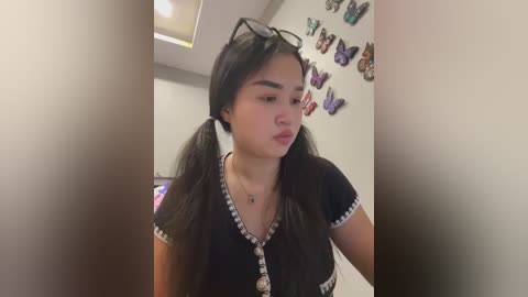 Media: Video of a young Asian woman with long black hair in pigtails, wearing a black V-neck top with white trim and butterfly earrings, standing in a room with butterfly wall art and butterfly decor.