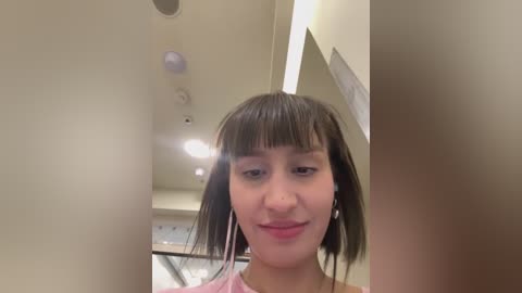 Media: Video of a young woman with straight, shoulder-length brown hair and bangs, wearing a pink shirt, standing in a modern, brightly lit hallway with beige walls and recessed lighting.