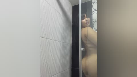 A video of a nude woman with light skin, medium-sized breasts, and a slim build, partially visible through a glass shower door in a modern bathroom with white and black tiled walls.