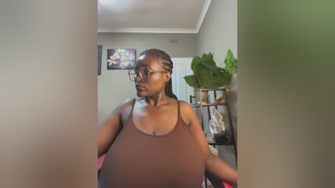 Media: A video of a Black woman with braided hair and glasses, wearing a brown tank top, standing in a dimly lit room with green plants and framed art on the wall.