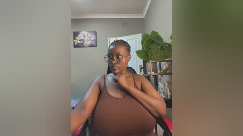 Media: A video of a pregnant woman with dark skin and glasses, wearing a brown tank top, sitting in a room with green walls, a potted plant, and a framed picture.