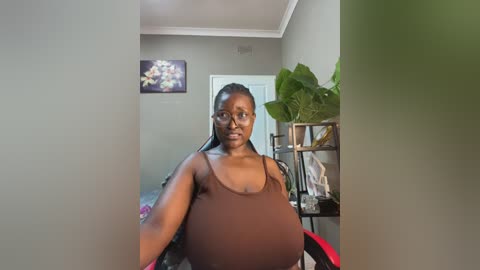 Media: Video of a pregnant woman with dark skin, long braids, and glasses, wearing a tight brown tank top, standing in a modern, beige-walled room with a large plant, framed art, and shelves.